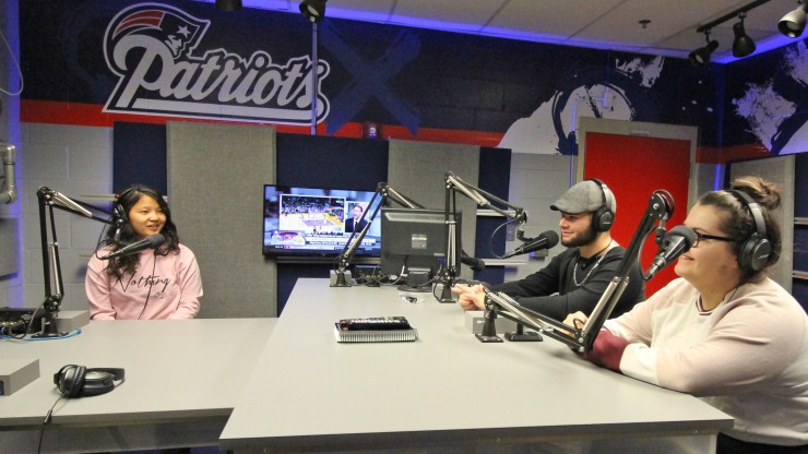 Three students sitting in front of microphones at the Patriots broadcasting station in Gillette Stadium.