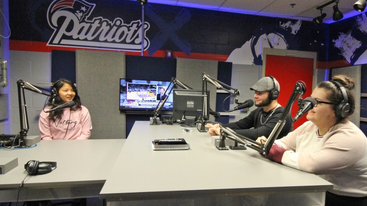 Three students wear headsets in front of microphones in the radio broadcasting stadium at Gillette Stadium.