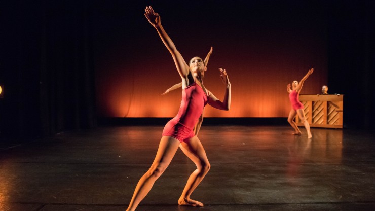 Three students on stage dancing with their arms pointed out during a performance on the Main Stage with a pianist playing in the background.