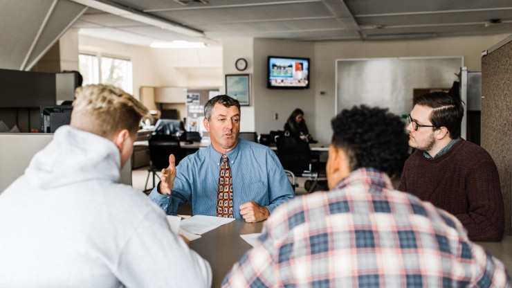 A professor sits with three students in an office to help them with classwork. 