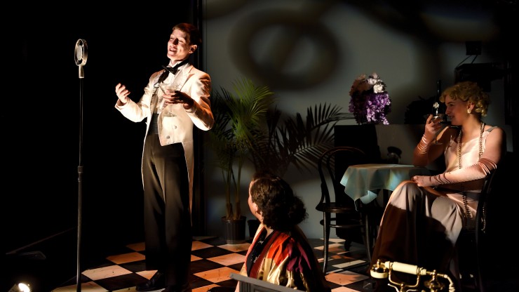 A student singing during a theatre performance while a student sits behind him in a chair and another student sits on the ground looking up.