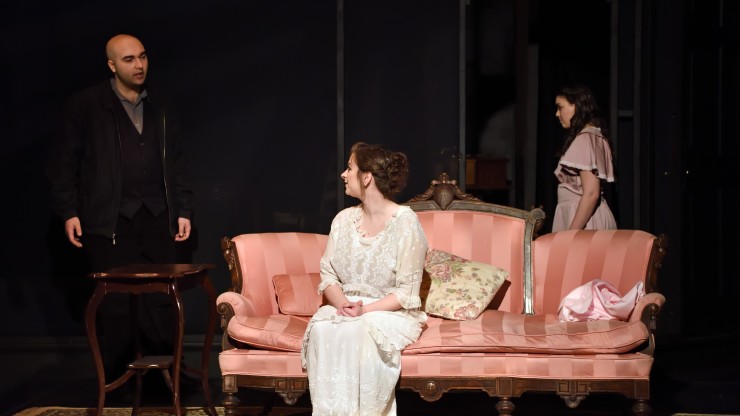A scene from a theatre performance. Two actors speaking to each other, one is standing and the other is sitting on a couch.